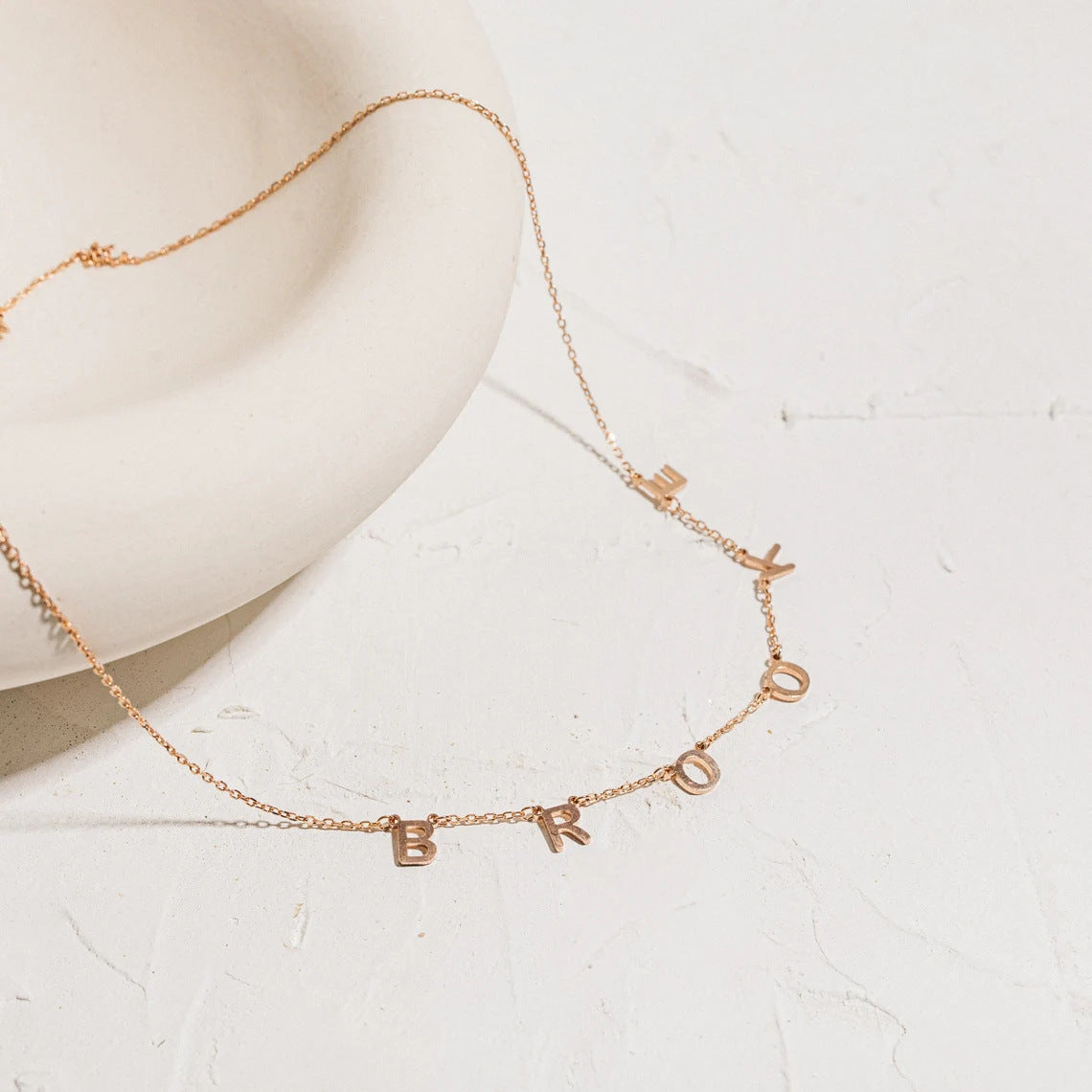 Amara initial necklace displayed on a white dish in rose gold with initials B R O O K E.